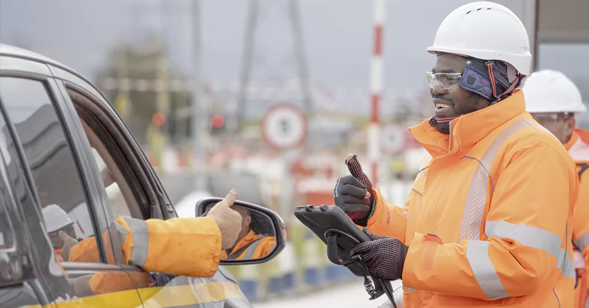 Construction site worker in high-visibility clothing and a hard hat checking a vehicle at a site entrance using a handheld device.