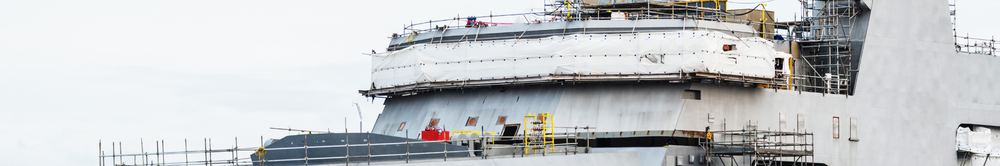 Close-up of a warship's superstructure under construction in a shipyard, surrounded by scaffolding, with visible steel plating and hull sections mid-assembly.