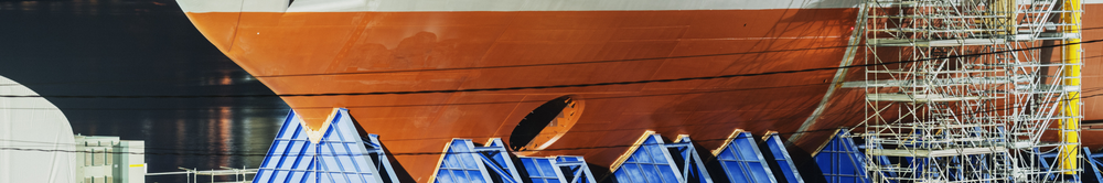 The orange hull of a ship under repair in a UK shipyard dry dock, with scaffolding rigged up one side and blue staging platforms along the waterline
