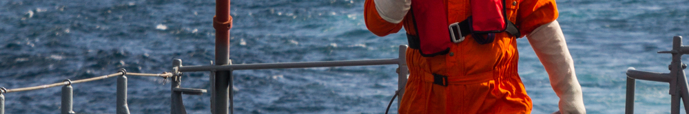 Worker in orange overalls and a red life jacket standing on a vessel's deck beside railings, with the sea behind